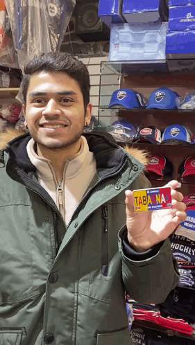 A Man In A Green Parka Holds Up A Card That Says Tabana GIF