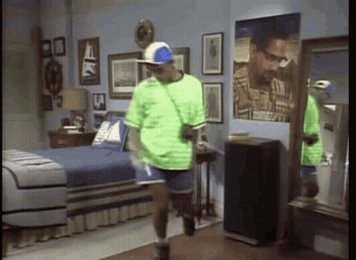 A Man In A Green Shirt And Blue Shorts Is Standing In A Bedroom . GIF