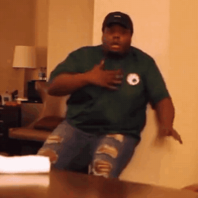 A Man In A Green Shirt And Ripped Jeans Is Standing In Front Of A Table . GIF