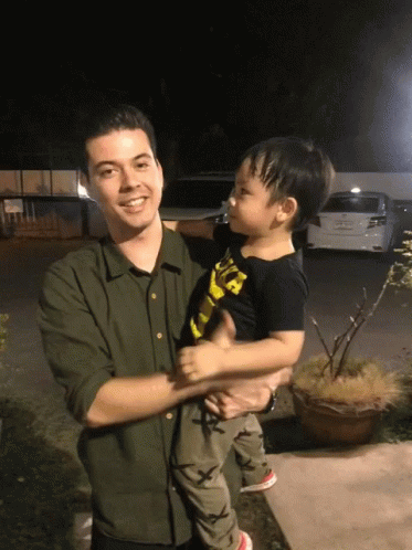A Man In A Green Shirt Holds A Little Boy In A Black Shirt That Says ' A ' On It GIF