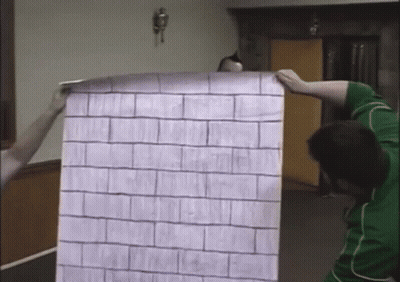 A Man In A Green Shirt Is Holding A Large Piece Of Paper With A Brick Pattern On It GIF