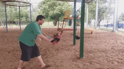 A Man In A Green Shirt Is Pushing A Child On A Swing At A Playground GIF