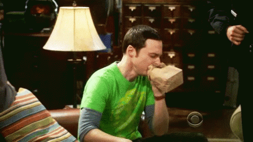 A Man In A Green Shirt Is Sitting On A Couch Holding A Paper Bag In His Mouth . GIF
