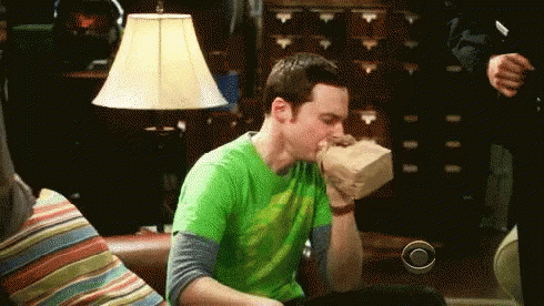 A Man In A Green Shirt Is Sitting On A Couch With A Bag In His Mouth . GIF