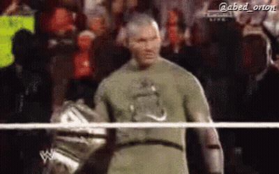 A Man In A Green Shirt Is Standing In A Wrestling Ring With A Crowd Behind Him . GIF