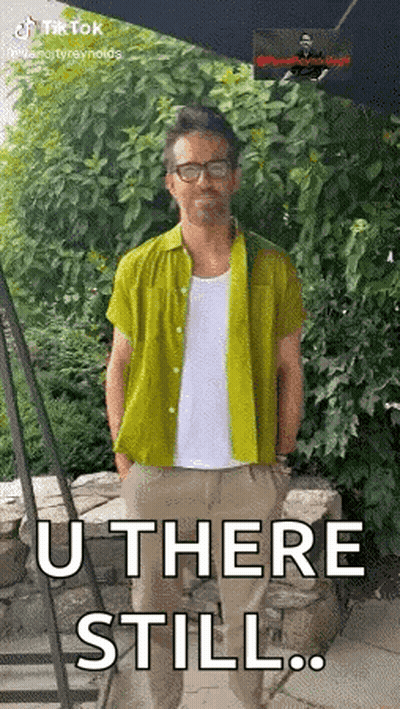 A Man In A Green Shirt Is Standing In Front Of A Stone Wall With His Hands In His Pockets . GIF