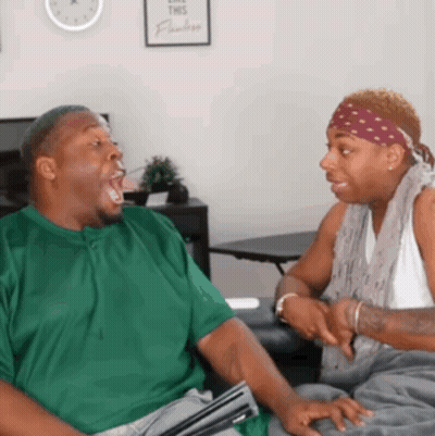A Man In A Green Shirt Is Talking To Another Man In A White Tank Top GIF
