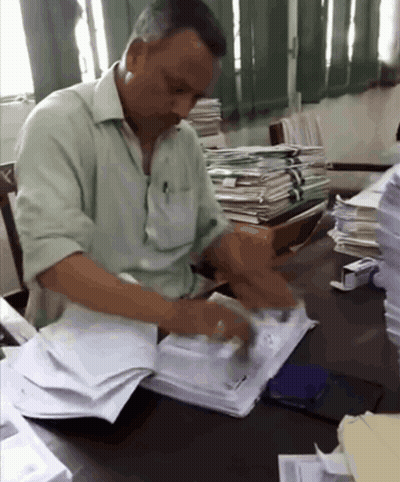 A Man In A Green Shirt Sits At A Desk With A Stack Of Papers GIF