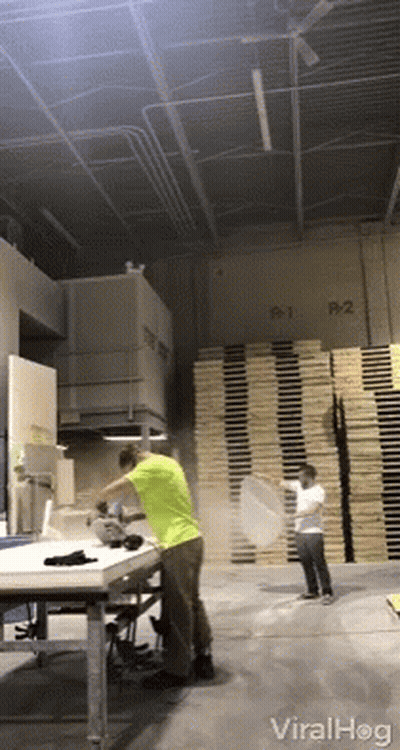 A Man In A Green Shirt Throws A Pizza In A Warehouse With A Stack Of Wooden Pallets In The Background GIF