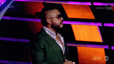 A Man In A Green Suit And Sunglasses Stands In Front Of A Purple And Orange Background That Says Aew On It GIF