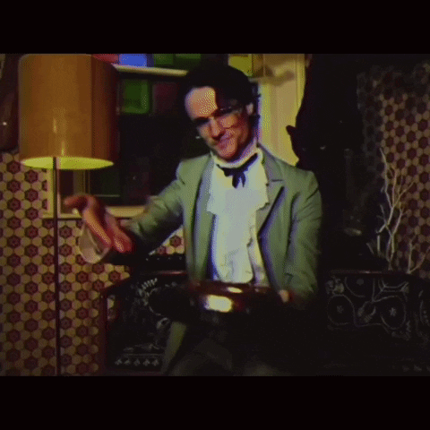 A Man In A Green Suit Is Playing A Drum In A Room GIF