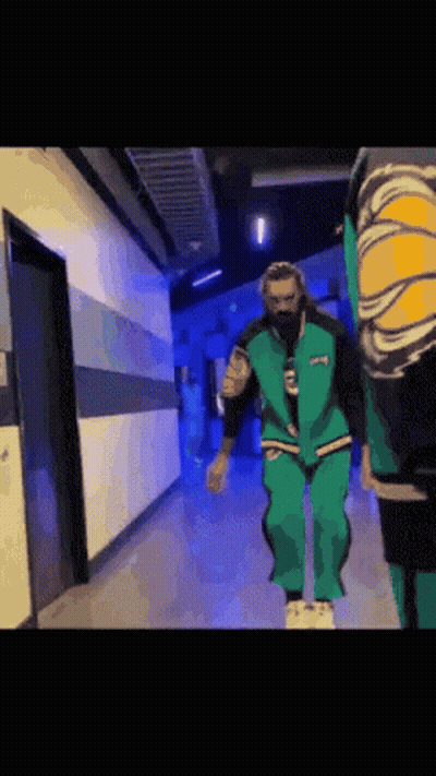 A Man In A Green Suit Is Walking Down A Hallway . GIF