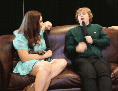 A Man In A Green Sweater Holds A Microphone While Sitting Next To A Woman GIF