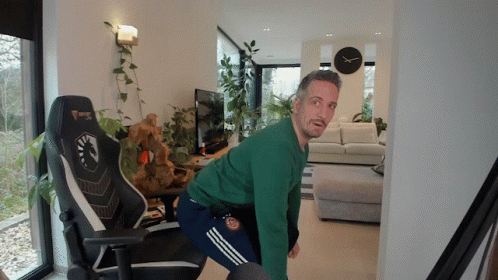 A Man In A Green Sweater Is Squatting In Front Of A Gaming Chair GIF