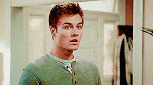 A Man In A Green Sweater Is Standing In Front Of A Door Looking Surprised . GIF