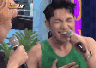 A Man In A Green Tank Top Is Singing Into A Microphone While Another Man Looks On .. GIF