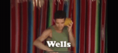 A Man In A Green Tank Top Is Standing In Front Of A Colorful Curtain Holding A Yellow Object In His Hand . GIF