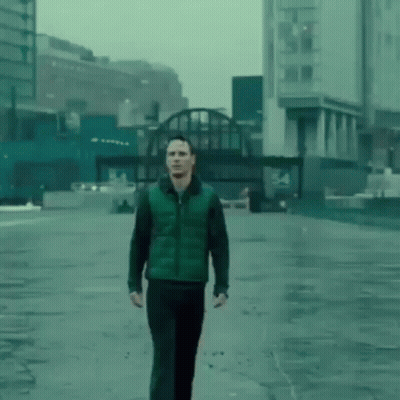 A Man In A Green Vest And Black Pants Is Walking Down A Wet Street In A City . GIF