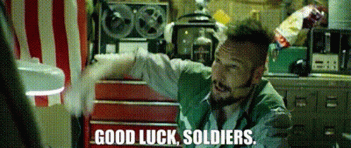 A Man In A Green Vest Says Good Luck Soldiers In Front Of A Red And White Striped Wall GIF
