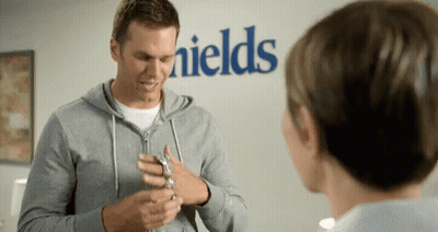 A Man In A Grey Hoodie Is Talking To A Woman In Front Of A Sign That Says Fields GIF