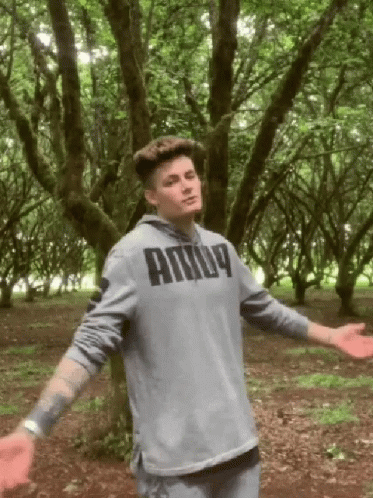 A Man In A Grey Puma Hoodie Stands In A Forest With His Arms Outstretched GIF