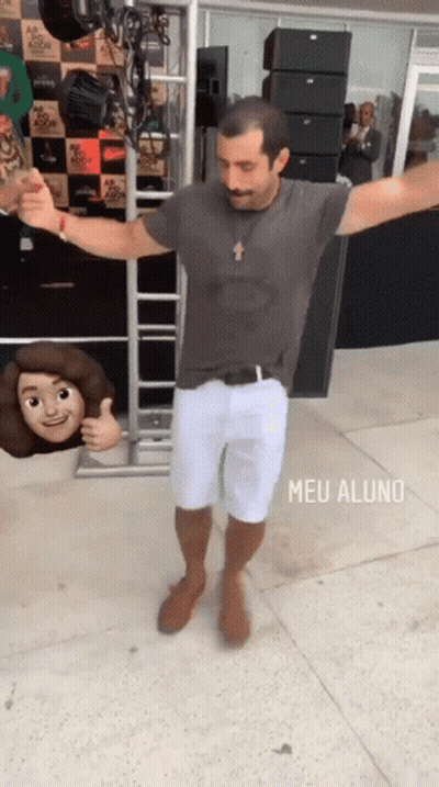 A Man In A Grey Shirt And White Shorts Is Dancing In Front Of A Sign That Says Meu Aluno On It GIF