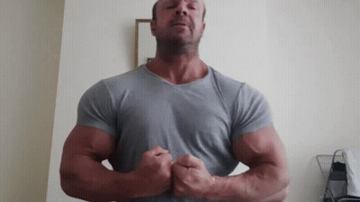 A Man In A Grey Shirt Is Flexing His Muscles In Front Of A White Wall GIF