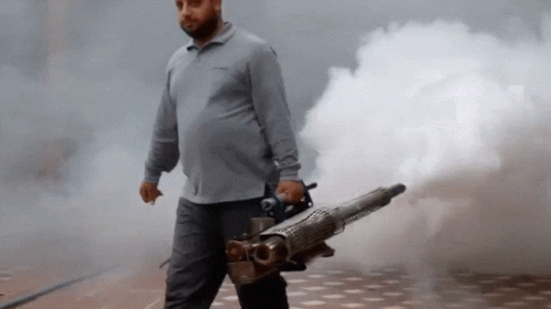 A Man In A Grey Shirt Is Holding A Machine That Is Shooting Smoke GIF