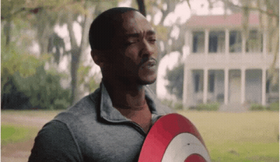 A Man In A Grey Shirt Is Holding A Red And White Shield In Front Of A House . GIF
