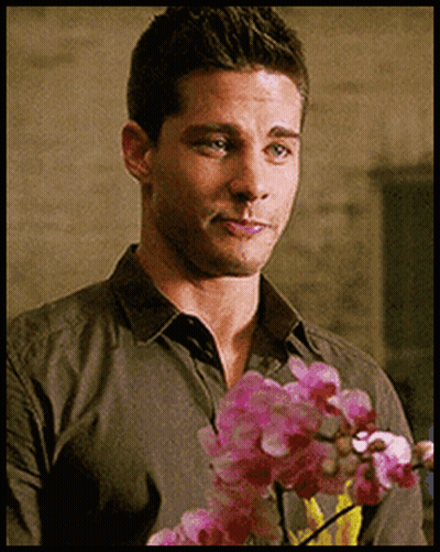 A Man In A Grey Shirt Is Holding Pink Flowers In His Hand GIF