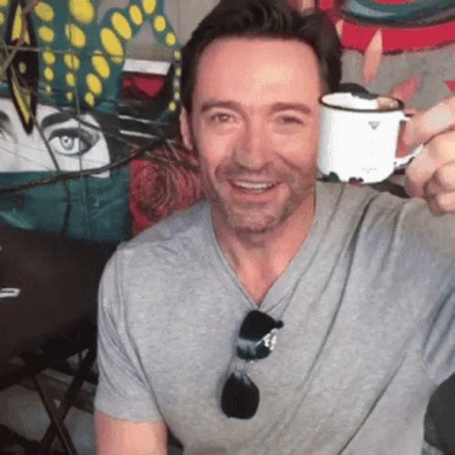 A Man In A Grey Shirt Is Smiling And Holding A White Cup GIF