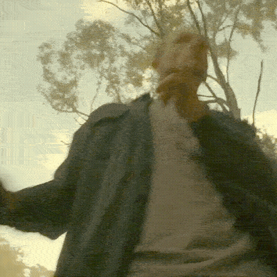A Man In A Grey Shirt Is Standing In Front Of A Tree GIF