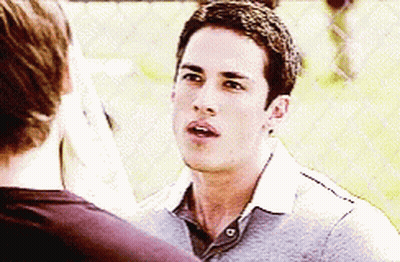A Man In A Grey Shirt Is Talking To Another Man Behind A Chain Link Fence GIF