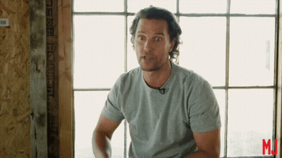 A Man In A Grey Shirt Stands In Front Of A Window With The Letter M On The Bottom GIF