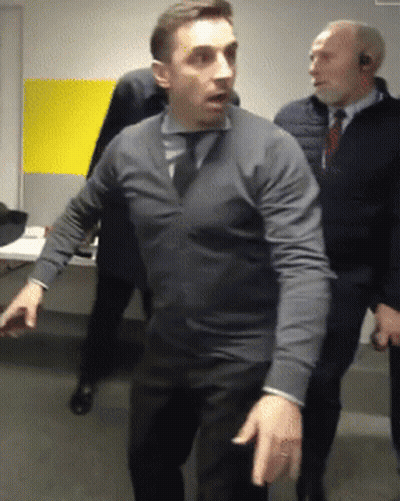 A Man In A Grey Sweater And Tie Stands In A Room With Other Men GIF