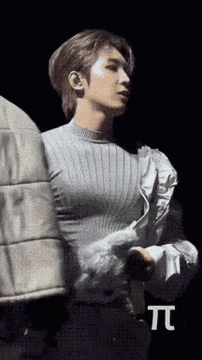 A Man In A Grey Sweater Is Standing In Front Of A Black Background With The Letter T. GIF