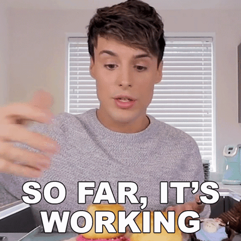 A Man In A Grey Sweater Says So Far It 'S Working In Front Of A Window GIF