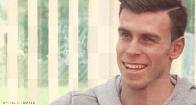 A Man In A Grey Sweatshirt Is Smiling In Front Of A Window With A Striped Blind Behind Him GIF