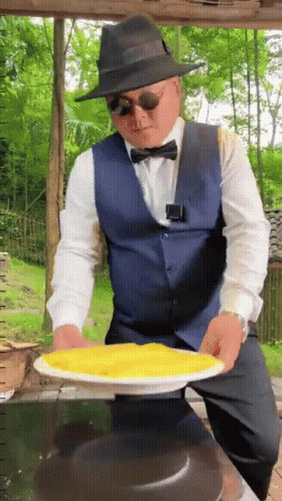 A Man In A Hat And Bow Tie Is Holding A Plate Of Food GIF