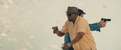 A Man In A Hat And Sunglasses Is Holding Two Guns In Front Of A Woman . GIF