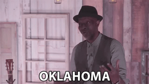 A Man In A Hat And Vest Is Standing In Front Of A Window And Holding A Guitar . GIF