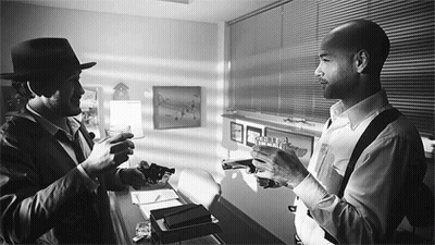 A Man In A Hat Holds A Gun Next To Another Man Who Holds A Gun GIF