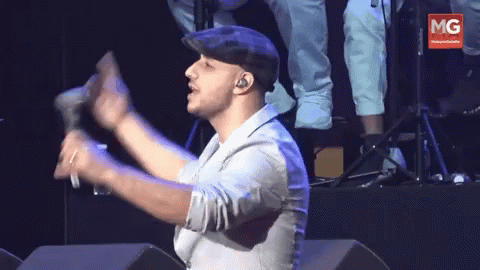 A Man In A Hat Holds A Microphone In Front Of A Banner That Says Mg GIF