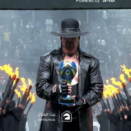 A Man In A Hat Holds A Trophy In Front Of Flames And A Sign That Says Baytal Hilal GIF
