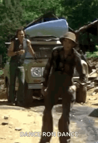 A Man In A Hat Is Dancing In Front Of A Truck With A Kayak On Top Of It . GIF
