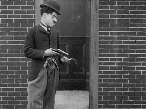 A Man In A Hat Is Holding A Gun In Front Of A Brick Building GIF