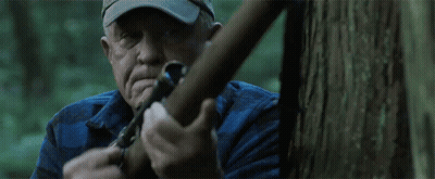 A Man In A Hat Is Holding A Sniper Rifle In Front Of A Tree GIF