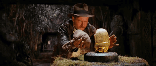 A Man In A Hat Is Looking At A Gold Object In His Hands GIF