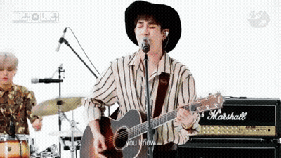 A Man In A Hat Is Playing An Acoustic Guitar In Front Of A Microphone . GIF