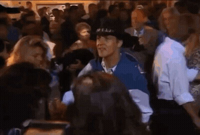A Man In A Hat Is Standing In A Crowd Of People . GIF
